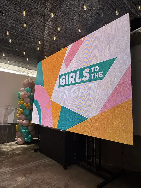 Pantalla con el logotipo de Girls To The Front en el patio del evento, junto al podio de acrílico y los globos.