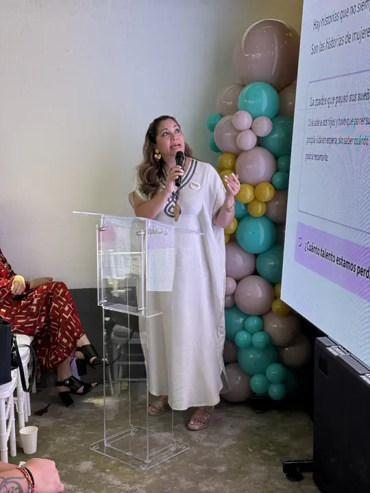 Ponente con vestido de lino blanco se dirige al público y mira hacia la pantalla.