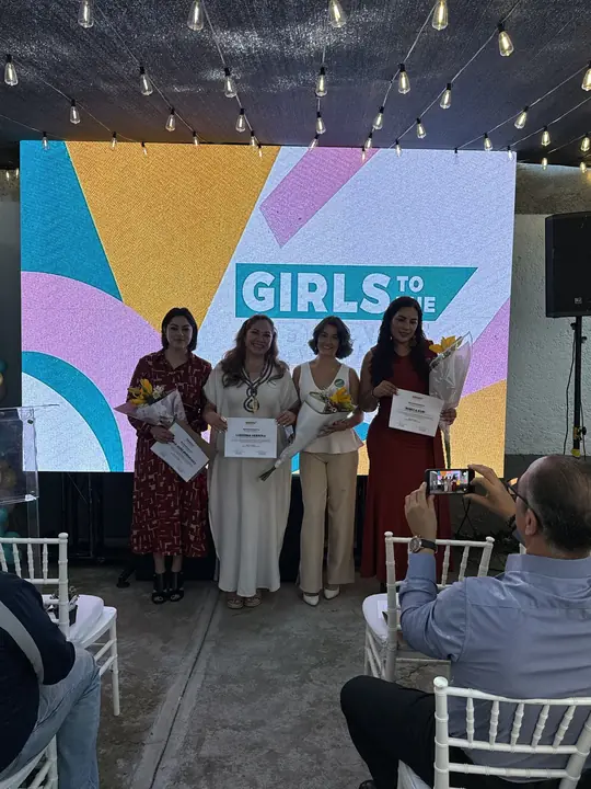 Cuatro mujeres sostienen reconocimientos y flores frente a la pantalla de Girls To The Front.