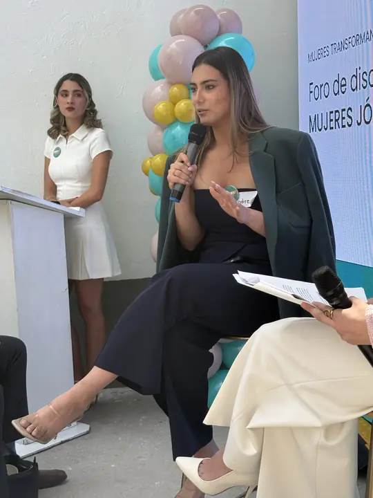 Conversación en formato panel: una participante habla con micrófono mientras la conductora escucha de pie junto al podio.
