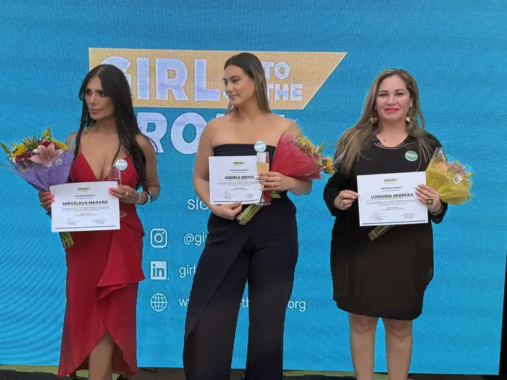Tres participantes sostienen reconocimientos y flores frente a la pantalla de Girls To The Front.